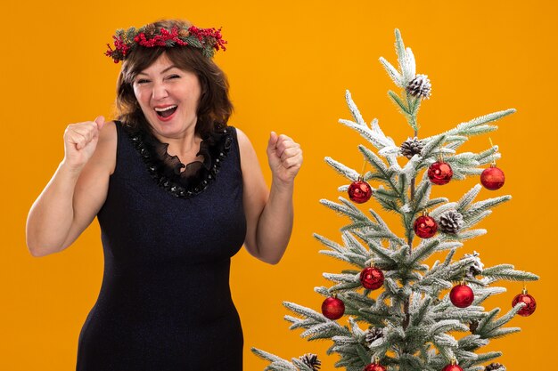 Joyful middle-aged woman wearing christmas head wreath and tinsel garland around neck standing near decorated christmas tree