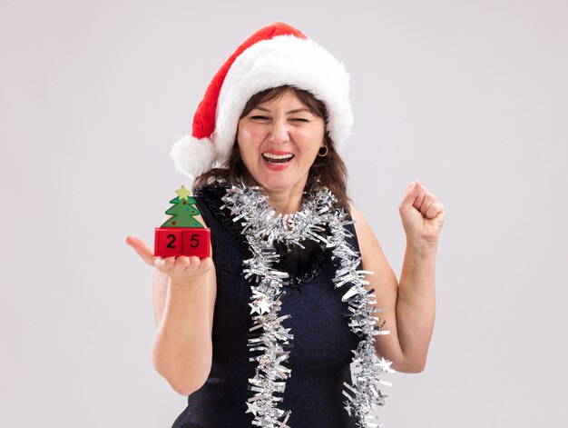 Joyful middle-aged woman wearing santa hat and tinsel garland around neck holding christmas tree toy with date looking at camera doing yes gesture isolated on white background