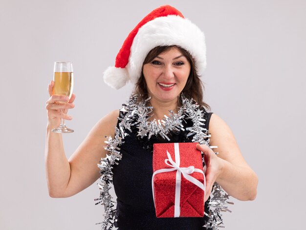 Joyful middle-aged woman wearing santa hat and tinsel garland around neck holding glass of champagne stretching out gift package towards camera looking at camera isolated on white background