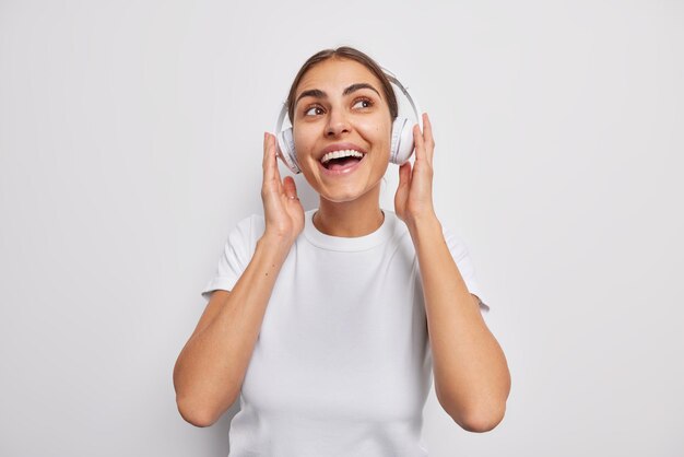 Joyful millennial girl listens favorite audio track in wireless headphones smiles broadly has happy expression dressed casually isolated over white wall