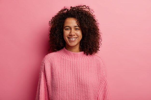 Радостная миллениальная девушка с густыми волосами афро зубасто улыбается, наслаждается счастливым моментом, с удовольствием смотрит в камеру, носит вязаный джемпер, изолированный на розовом фоне.
