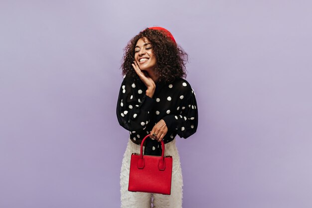 Joyful modern girl with wavy hairstyle and cool cap in polka dot black blouse smiling and holding red stylish handbag on lilac wall