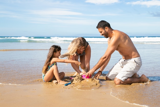 Joyful mom, dad and little daughter enjoying vacation at sea together, playing with daughters sand toys, building sandcastle
