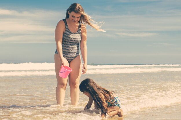 Радостная мама и дочка стоят по щиколотку в морской воде и мокром песке, собирая ракушки в ведро