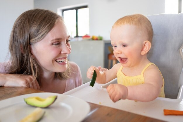 Радостная мама смотрит, как ребенок ест твердую пищу в высоком стульчике, смеется и веселится. Снимок крупным планом. Концепция ухода за детьми или питания
