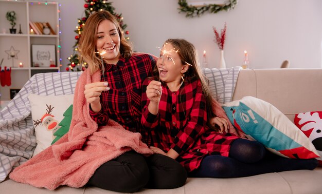 Joyful mother and daughter holding sparklers covered with blanket sitting on couch and enjoying christmas time at home