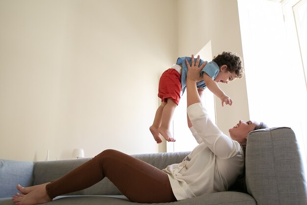 Joyful mother lying on sofa and holding son on straight hands. Funny mix-raced boy having fun with caring mom. Smiling mum playing with toddler and rising his up. Family and motherhood concept