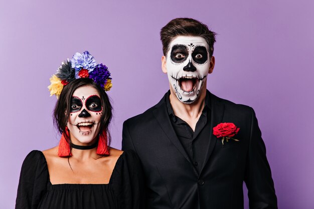 Joyful naughty men and woman in black clothes with red details scream in amazement, posing with Halloween makeup for portrait.