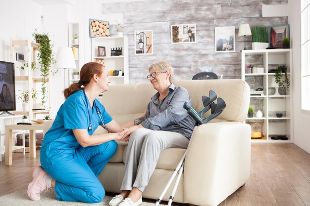 Joyful old lady sitting on couch in a nursing home holding health taker arm.