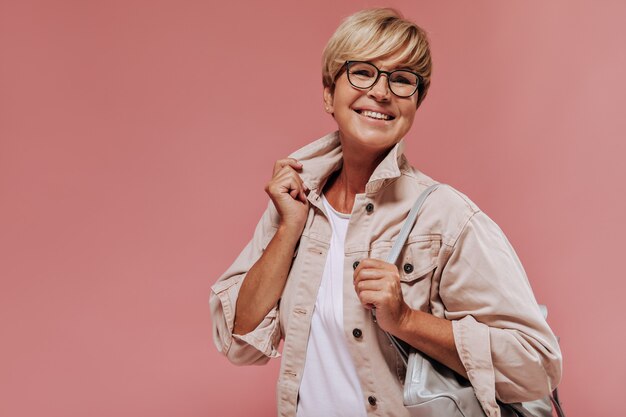 Joyful old lady with blonde short hairstyle and tanned skin in beige modern jacket smiling and holding cool bag on isolated backdrop. 