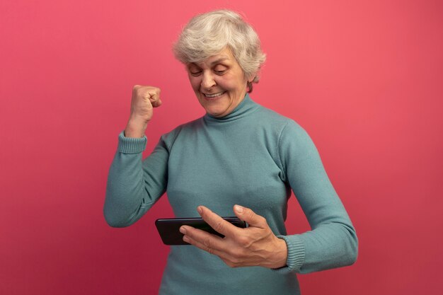 Joyful old woman wearing blue turtleneck sweater holding and looking at mobile phone doing yes gesture 