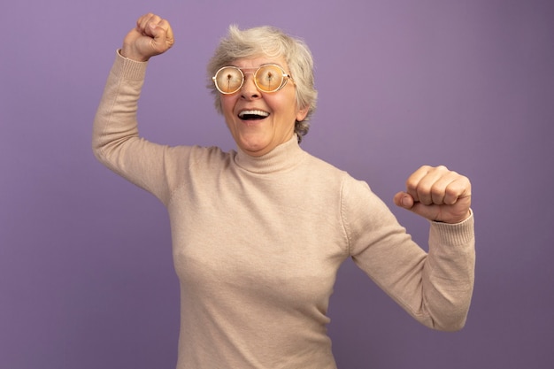 Free photo joyful old woman wearing creamy turtleneck sweater and glasses looking at side raising fists up isolated on purple wall