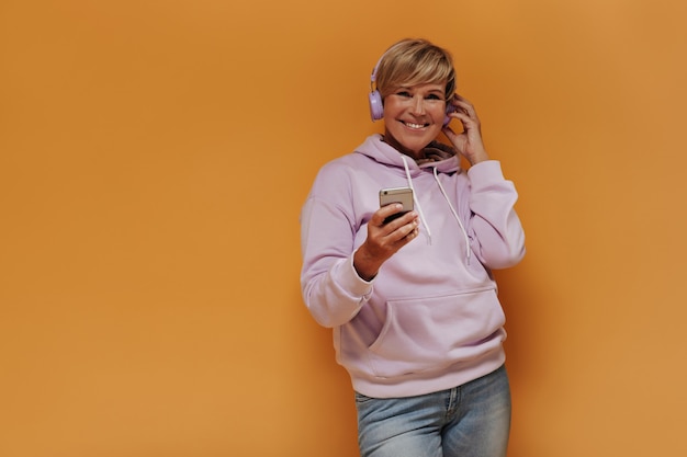 Joyful old woman with stylish blonde hairstyle and lilac headphones in trendy pink hoodie and jeans smiling and holding smartphones.