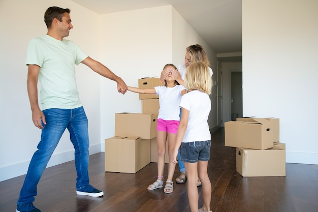 Joyful parents and little sister leading girl with closed eyes in new apartment