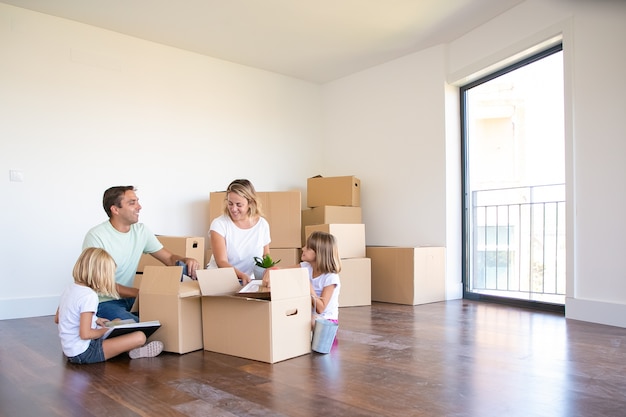 Радостные родители и двое детей распаковывают вещи в новой пустой квартире, сидят на полу и достают предметы из открытых ящиков