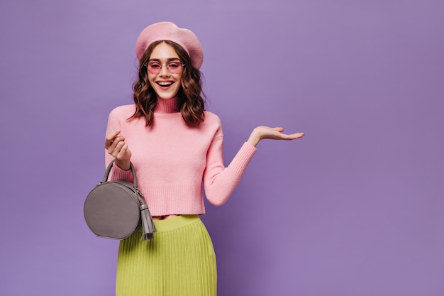 Joyful Parisian woman in beret and sunglasses points at place for text on purple wall