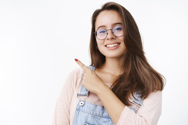 Joyful pleasant young female assistant in glasses and overalls smiling broadly pointing at upper left corner showing awesome place to hang out asking click on copy space over gray background