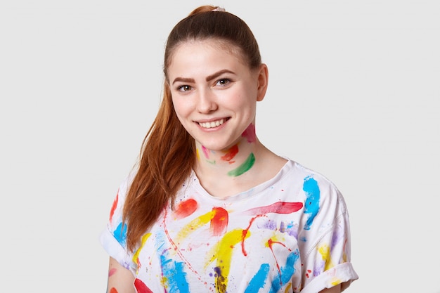 즐거운 유럽 여성 예술가는 포니 테일, 이빨 미소, 캐주얼 티셔츠를 입은 하얀 치아도 보여줍니다.