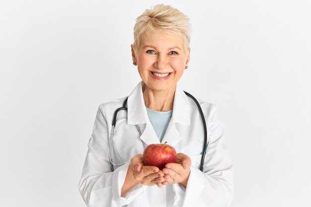 Joyful positive mature female practitioner holding sweet crunchy fruit rich in fiber, phytonutrients and antioxidants, recommending to eat healthy organic food. Apple a day keeps doctor away