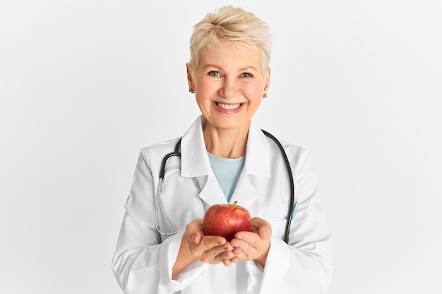 Бесплатное фото Радостная и позитивная зрелая практикующая женщина держит в руках сладкие хрустящие фрукты, богатые клетчаткой, фитонутриентами и антиоксидантами, и рекомендует есть здоровую органическую пищу. яблоко в день держит доктора подальше