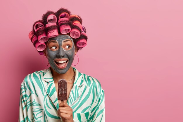 Joyful positive woman wears hair rollers for curls and beauty mask, eats delicious chocolate ice cream, wears casual robe, looks aside happily, isolated on pink , blank copy space.