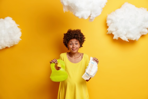 Joyful pregnant woman has Afro hair, holds diaper and rubber bib for baby, dressed in casual dress, prepares for child birth, buys all necessary things for newborn, isolated on yellow .