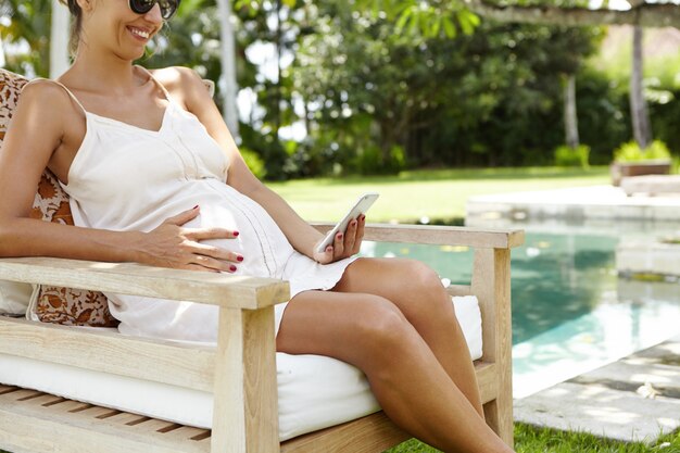 ヘルスリゾートでの休暇中に屋外のベンチで休んでいる間彼女の携帯電話でメッセージを読んで幸せな笑顔でうれしそうな妊娠中の女性