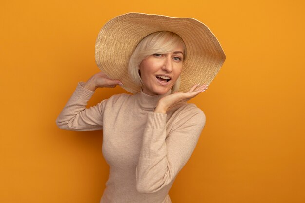 Joyful pretty blonde slavic woman with beach hat holds hand open isolated on orange wall