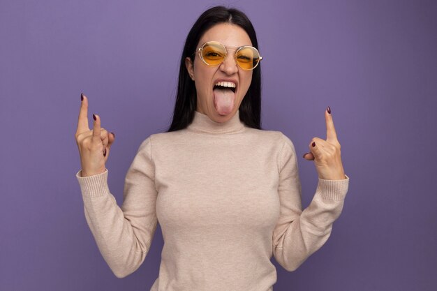 Joyful pretty brunette caucasian girl in sun glasses stucks out tongue and gestures horns with two hands on purple