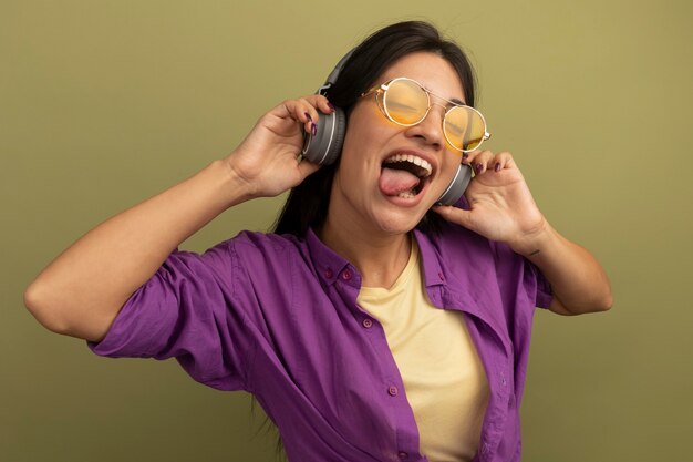 Joyful pretty brunette caucasian girl in sun glasses with headphones stucks out tongue on olive green