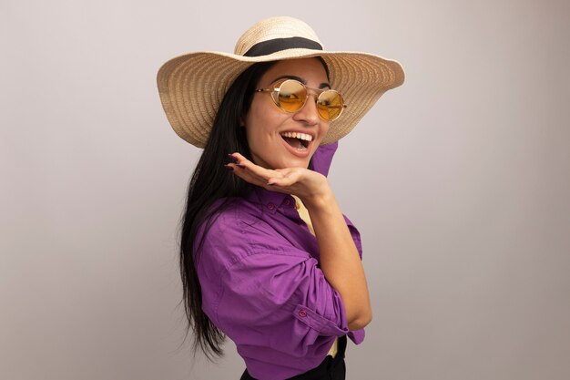 Joyful pretty brunette woman in sun glasses with beach hat stands sideways puts hand on chin isolated on white wall