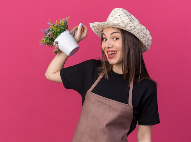 Joyful pretty caucasian female gardener wearing gardening hat holding flowerpot on pink