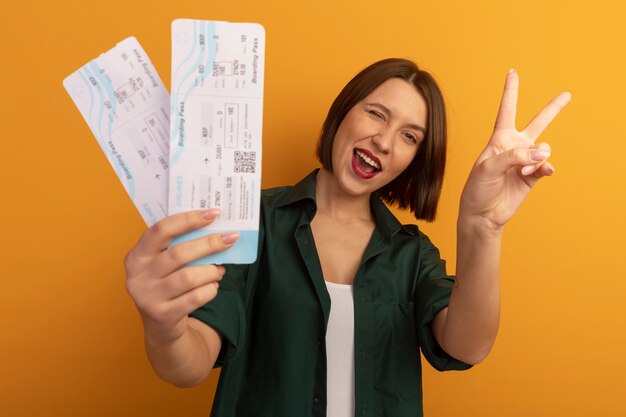 Joyful pretty caucasian woman blinks eye and gestures victory hand sign holding air tickets on orange