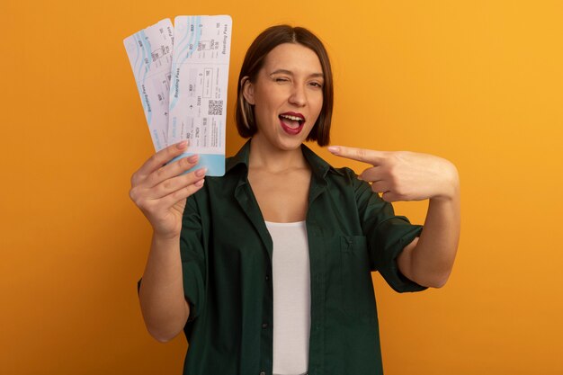 joyful pretty caucasian woman blinks eye holding and pointing at air tickets on orange