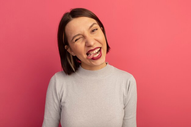 Joyful pretty caucasian woman blinks eye and stucks out tongue looking at camera on pink