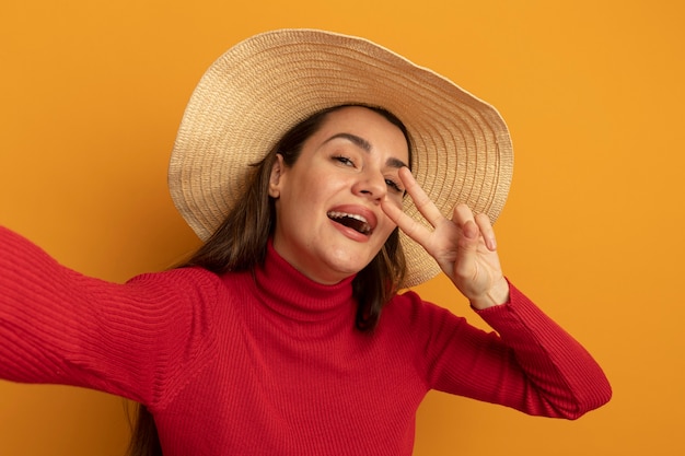 Joyful pretty caucasian woman with beach hat gestures victory hand sign and pretends to hold camera on orange