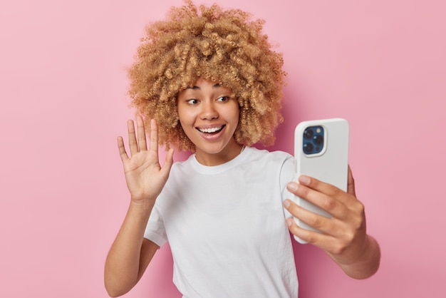 Joyful pretty curly haired Teenage girl waves hello at front camera of smartphone talks by video call greets friend on distance wears casual white t shirt isolated over pink background Technology