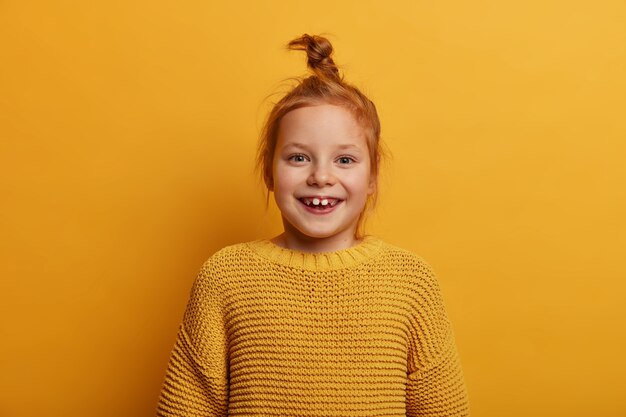 Joyful pretty little child with natural ginger hair, smiles positively, looks  with positive mood, wears knitted yellow sweater, notices something incredible and appealing, smiles toothily