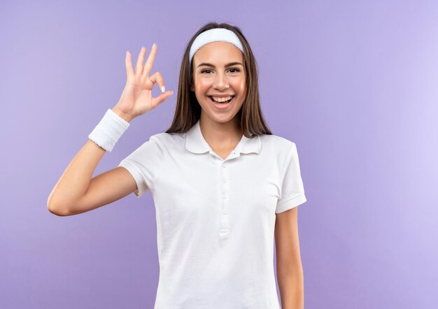 Joyful pretty sporty girl wearing headband and wristband doing ok sign isolated on purple wall with copy space