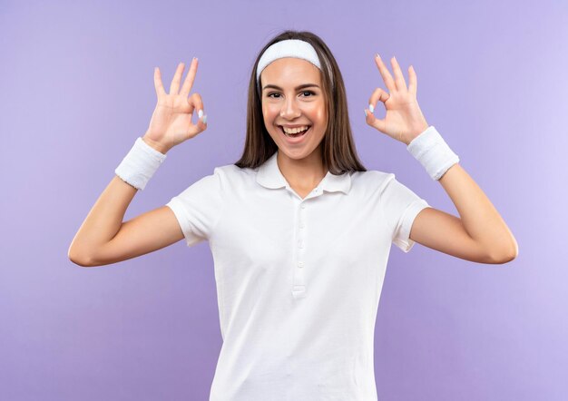 Joyful pretty sporty girl wearing headband and wristband doing ok signs isolated on purple wall