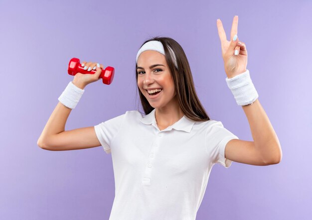 Joyful pretty sporty girl wearing headband and wristband holding dumbbell doing peace sign isolated on purple wall