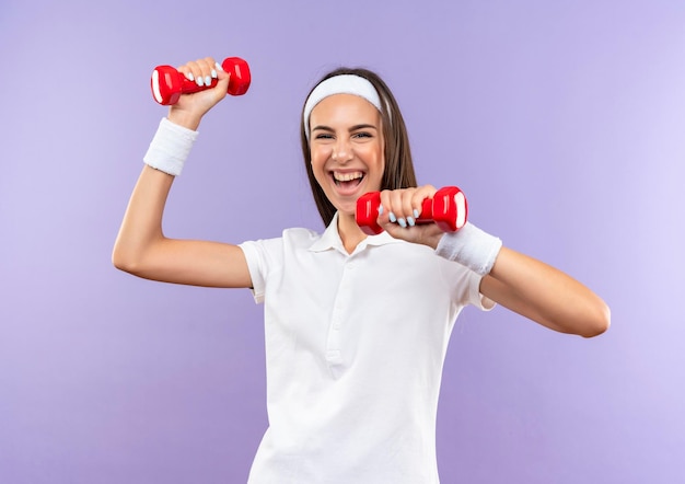 Joyful pretty sporty girl wearing headband and wristband holding dumbbells isolated on purple wall