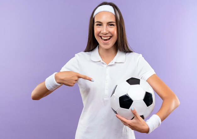 Joyful pretty sporty girl wearing headband and wristband holding and pointing at soccer ball isolated on purple wall