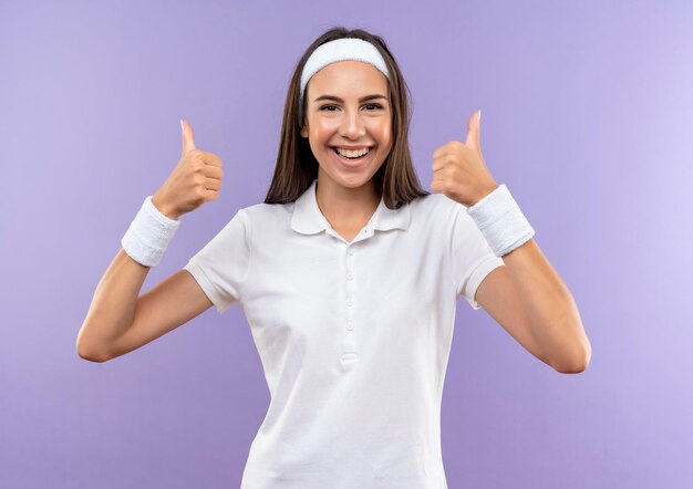 Радостная симпатичная спортивная девушка с повязкой на голову и браслет показывает большие пальцы руки вверх изолированной на фиолетовой стене
