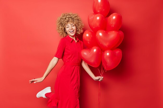 Joyful pretty woman dances with heart balloons enjoys celebration and party wears long dress smiles broadly expresses positive emotions isolated over red background Happy Valentines Day concept