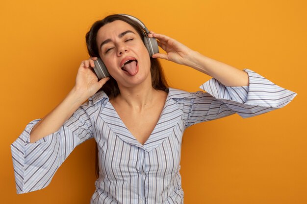 Joyful pretty woman on headphones blinks eye and stucks out tongue isolated on orange wall