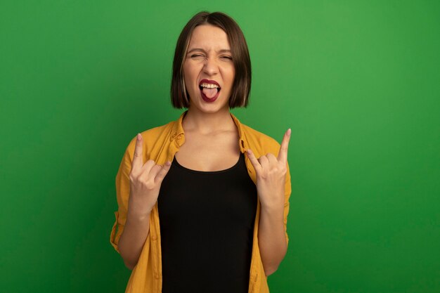 Joyful pretty woman stucks out tongue and gestures horns hand sign with two hands isolated on green wall