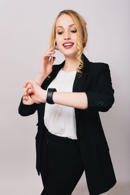 Joyful pretty young businesswoman in office suit talking on phone, smiling and looking at watch. Cheerful mood, happy, successful, worker, isolated, corporate