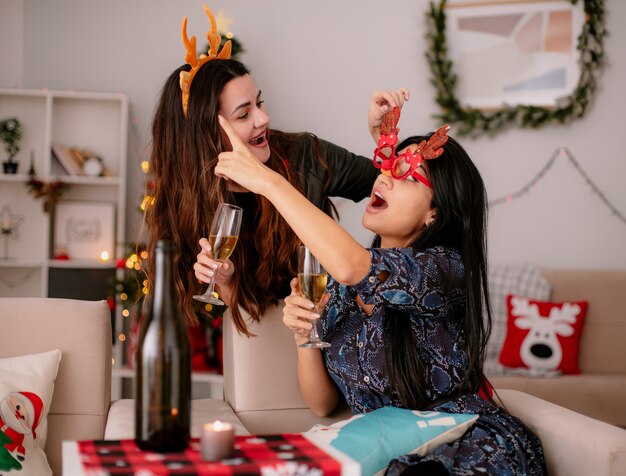 Joyful pretty young girl with reindeer headband holds glass of champagne and looks at her friend with reindeer glasses pointing at side sitting on armchair and enjoying christmas time at home