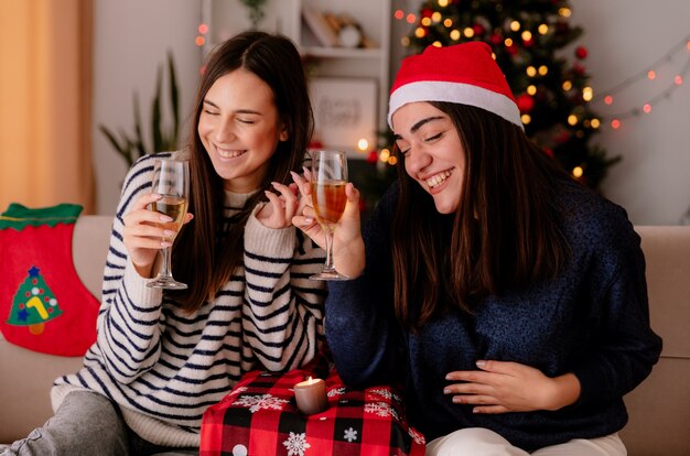 Joyful pretty young girls hold glasses of champagne sitting on armchairs and enjoying christmas time at home